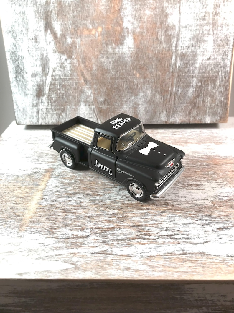 May include: A black toy truck with the words "Ring Bearer" on the roof and "Jimmy" on the side. The truck has a white bow tie design on the front and a striped bed.