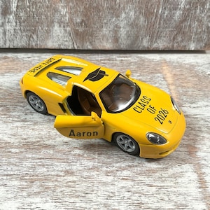 Puede incluir: Un coche de juguete amarillo con la puerta abierta, que muestra el nombre "Aaron". El coche tiene "CLASS OF 2026" impreso en el lateral y "DATE HERE" en el techo. Es una r&eacute;plica en miniatura.
