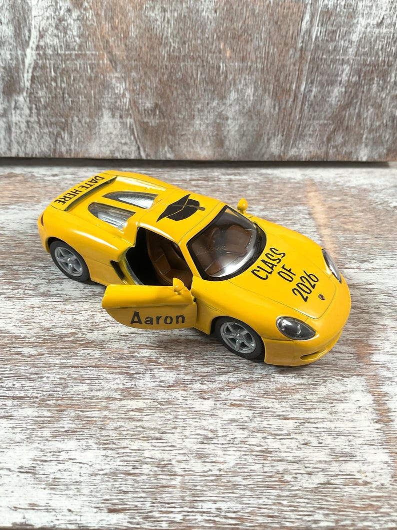 Puede incluir: Un coche de juguete amarillo con la puerta abierta, que muestra el nombre "Aaron". El coche tiene "CLASS OF 2026" impreso en el lateral y "DATE HERE" en el techo. Es una r&eacute;plica en miniatura.