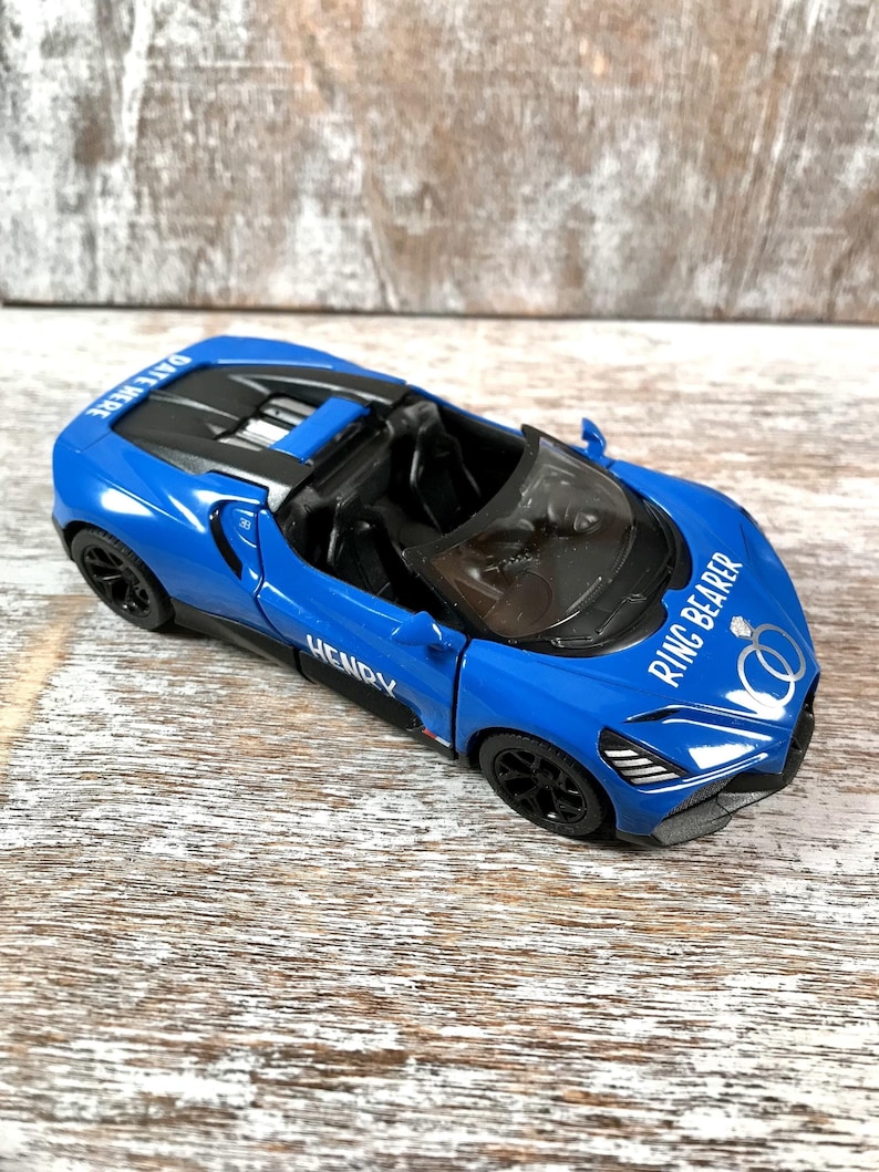 May include: A blue toy convertible car with black wheels. The car has the text "DATE HERE" on the top and "RING BEARER" with two wedding rings on the side. The name "HENRY" is also on the side.