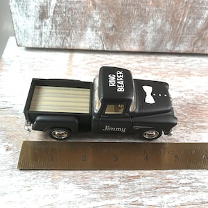 May include: A black toy truck with the words "Ring Bearer" on the roof and "Jimmy" on the side. The truck has a wooden bed and a white bow tie design. A ruler is placed next to the truck.