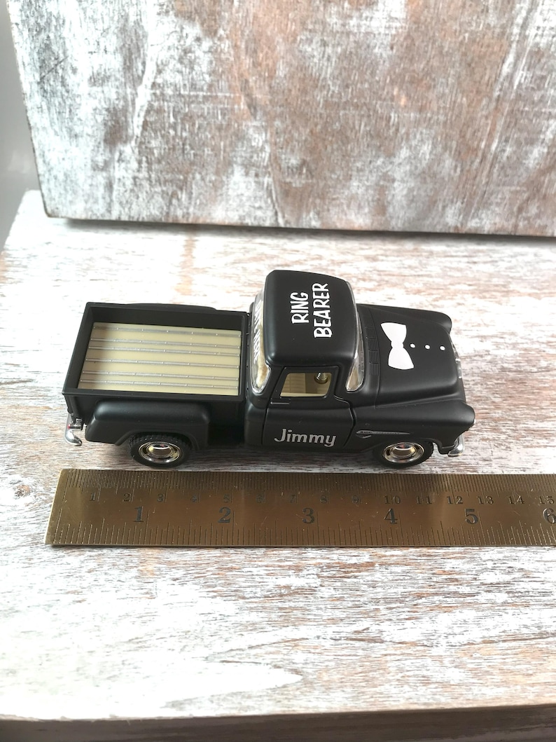 May include: A black toy truck with the words "Ring Bearer" on the roof and "Jimmy" on the side. The truck has a wooden bed and a white bow tie design. A ruler is placed next to the truck.