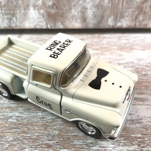 May include: A cream-colored toy pickup truck with the words "RING BEARER" on the roof and "Greg" on the door. The truck features a black bow tie graphic and three dots. The truck has a silver front bumper and wheels.