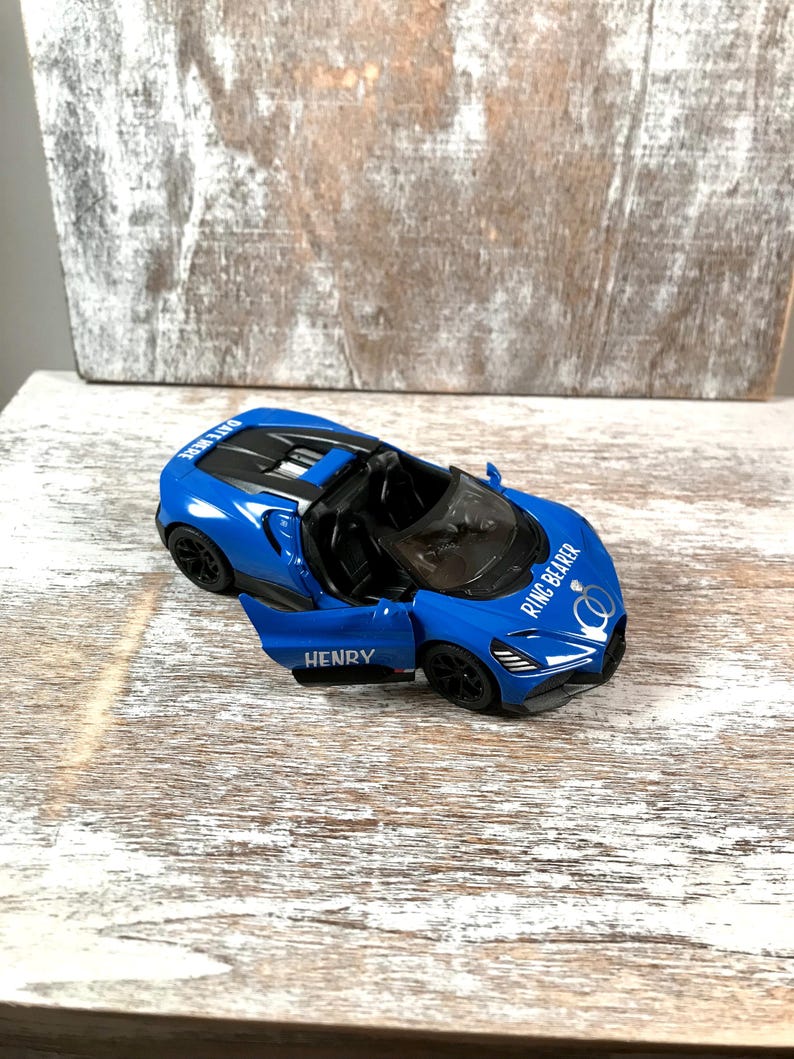 May include: A blue toy convertible car with the door open, displaying the name "HENRY". The car has black wheels and the text "RING BEARER" on the front. The car is on a wooden surface.
