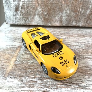 Puede incluir: Un coche de juguete amarillo con el texto "CLASS OF 2025" y "MAY 2025" impreso. El coche tiene detalles en negro y ruedas plateadas. El nombre "Thomas" tambi&eacute;n est&aacute; impreso en el coche.