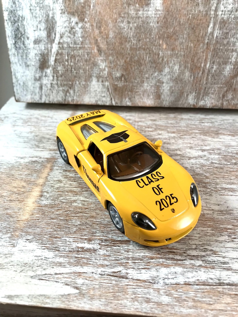 Puede incluir: Un coche de juguete amarillo con el texto "CLASS OF 2025" y "MAY 2025" impreso. El coche tiene detalles en negro y ruedas plateadas. El nombre "Thomas" tambi&eacute;n est&aacute; impreso en el coche.
