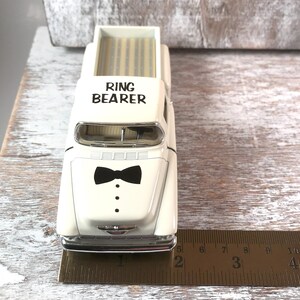 May include: A cream-colored toy pickup truck with the words "RING BEARER" printed on the cab. The front of the truck features a black bow tie and three black dots. The truck is approximately 4 inches long.