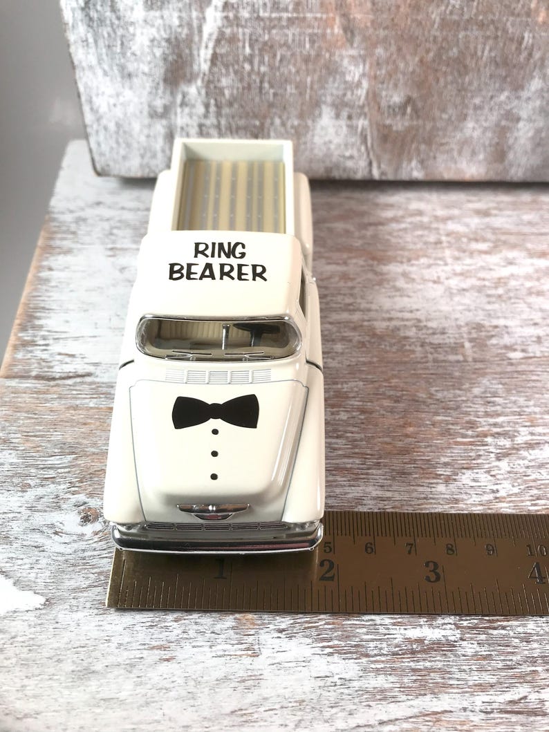 May include: A cream-colored toy pickup truck with the words "RING BEARER" printed on the cab. The front of the truck features a black bow tie and three black dots. The truck is approximately 4 inches long.