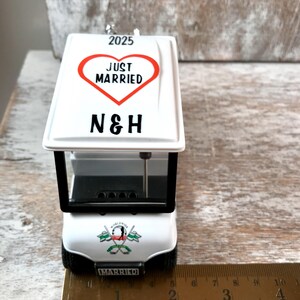 May include: White golf cart ornament with a red heart that says "Just Married" and the initials "N&H" on top. The cart has a black windshield and a "Worldwide Married" logo on the back. The ornament is 4 inches long.