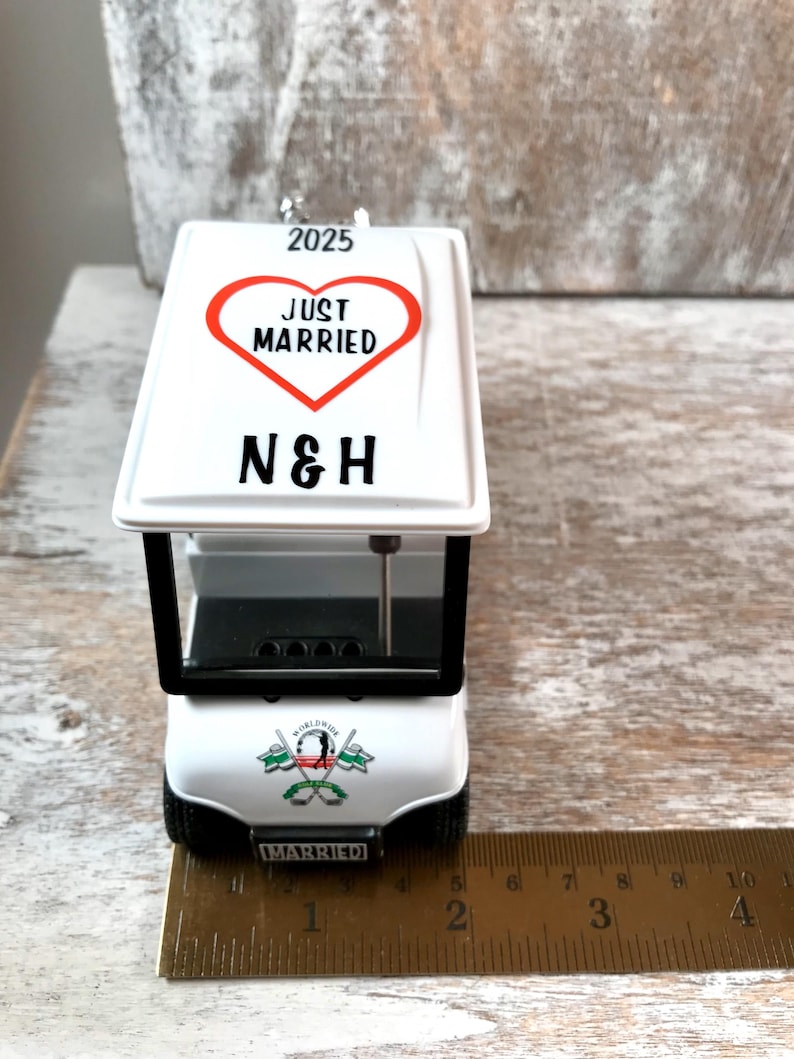 May include: White golf cart ornament with a red heart that says "Just Married" and the initials "N&H" on top. The cart has a black windshield and a "Worldwide Married" logo on the back. The ornament is 4 inches long.
