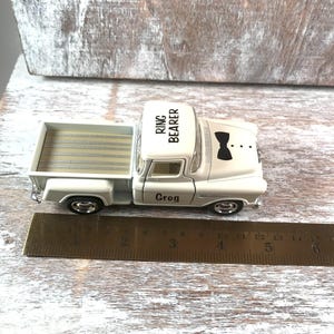 May include: A white toy pickup truck with the words "RING BEARER" on the roof and "Greg" on the door. The truck has a black bow tie graphic on the front. A ruler is next to the truck.