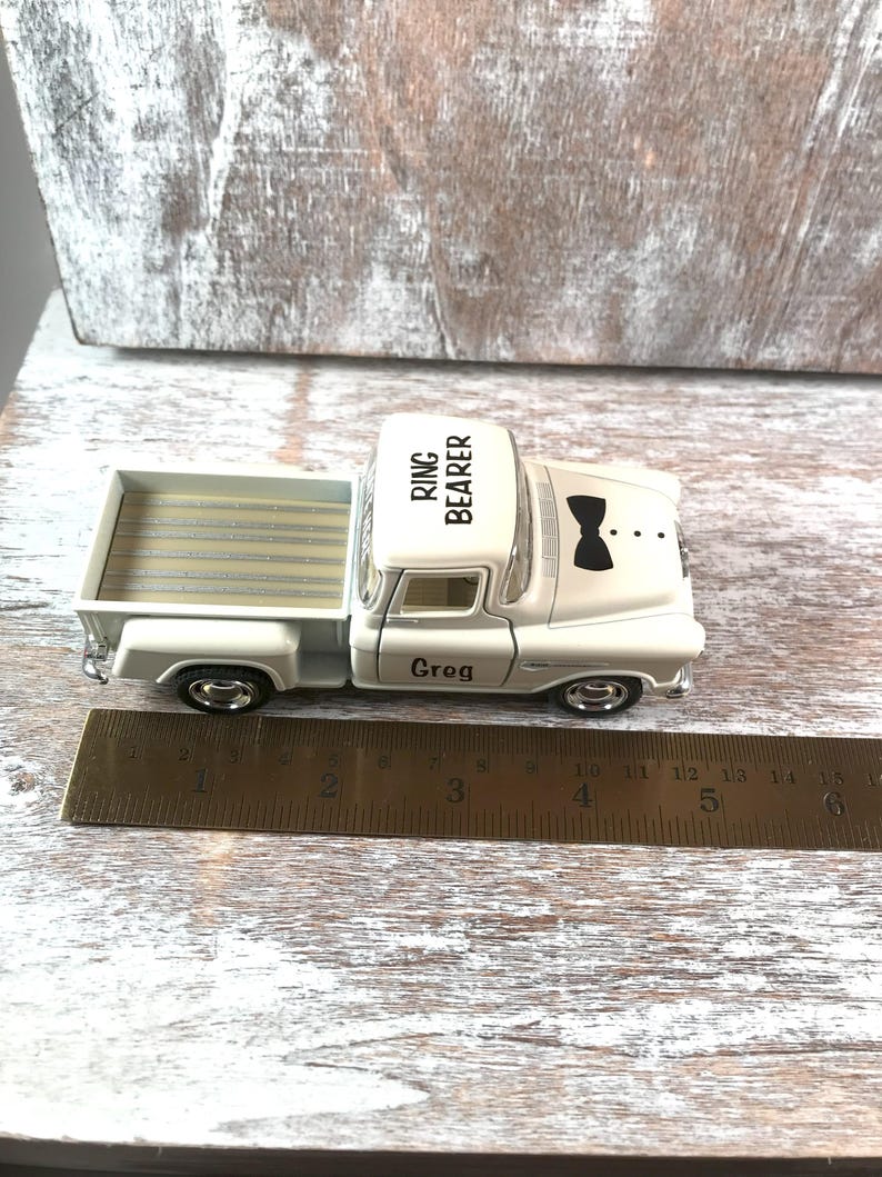 May include: A white toy pickup truck with the words "RING BEARER" on the roof and "Greg" on the door. The truck has a black bow tie graphic on the front. A ruler is next to the truck.