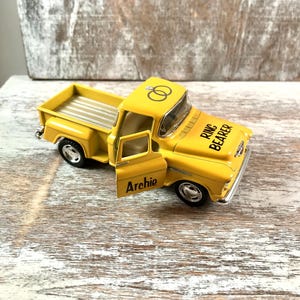 May include: A yellow toy pickup truck with the door open, featuring the text "Archie" on the door and "Ring Bearer" on the hood. The truck has a silver bed and two wedding rings on the roof.