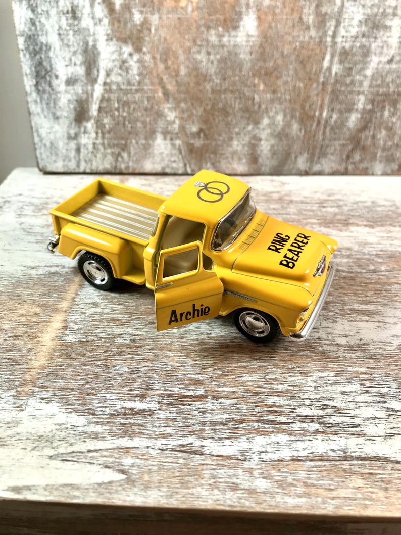 May include: A yellow toy pickup truck with the door open, featuring the text "Archie" on the door and "Ring Bearer" on the hood. The truck has a silver bed and two wedding rings on the roof.