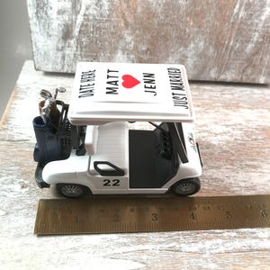 May include: A white golf cart figurine with black accents, a blue golf bag, and a roof that reads "MATT, JENN, JUST MARRIED" with a red heart. The cart has the number 22 on the side. A ruler shows the cart is approximately 6 inches long.