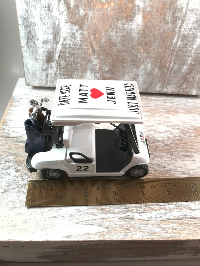 May include: A white golf cart figurine with black accents, a blue golf bag, and a roof that reads "MATT, JENN, JUST MARRIED" with a red heart. The cart has the number 22 on the side. A ruler shows the cart is approximately 6 inches long.