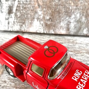 May include: A red toy truck with the words "Ring Bearer" and the name "Luke" in white lettering. The truck has a graphic of two wedding rings on the roof. The truck bed is tan and the wheels are black.