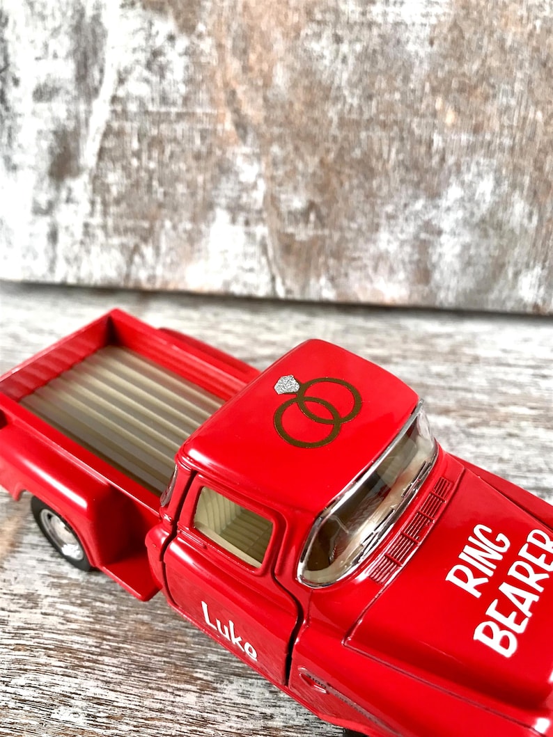May include: A red toy truck with the words "Ring Bearer" and the name "Luke" in white lettering. The truck has a graphic of two wedding rings on the roof. The truck bed is tan and the wheels are black.