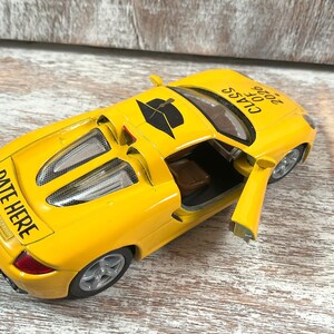 Puede incluir: Un coche de juguete amarillo brillante con la puerta abierta, mostrando el interior. El coche tiene el texto "CLASS OF 2026" en el techo y "DATE HERE" en la parte trasera. El coche tiene ruedas plateadas.
