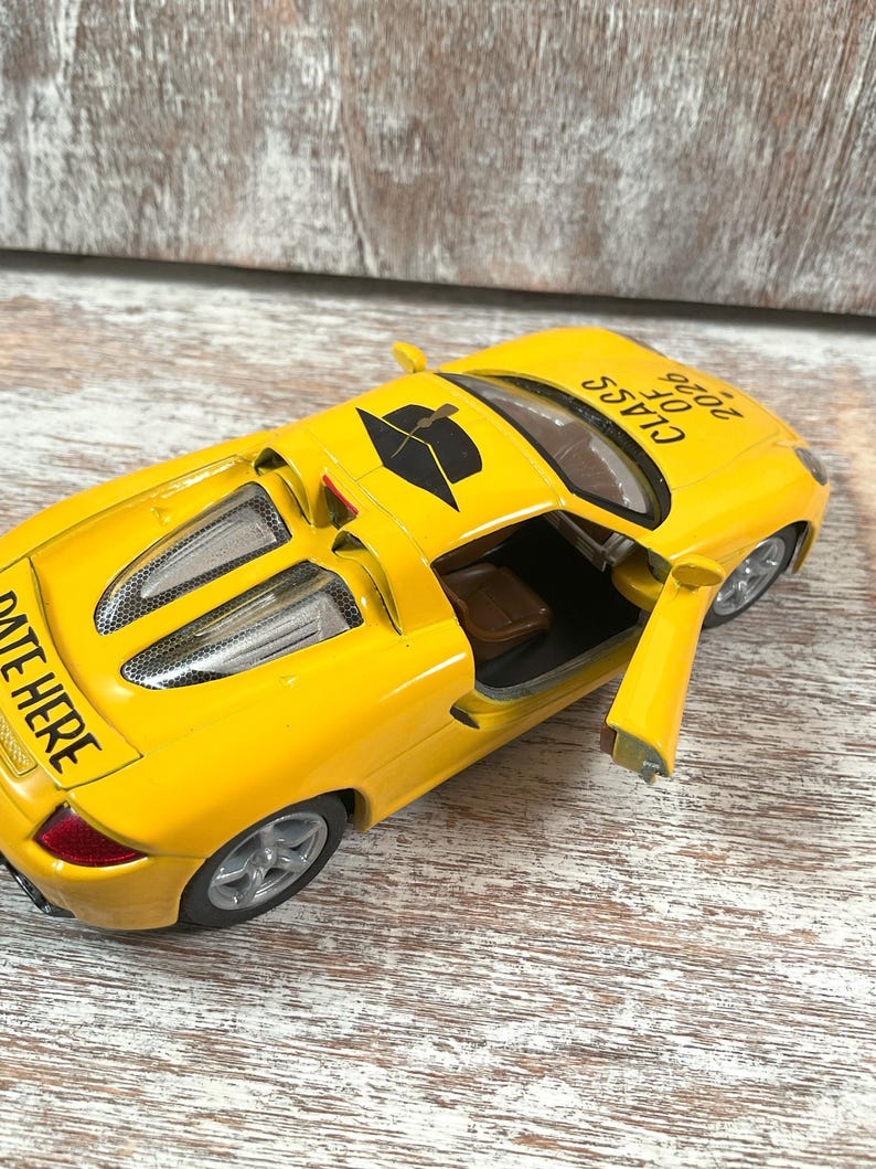 Puede incluir: Un coche de juguete amarillo brillante con la puerta abierta, mostrando el interior. El coche tiene el texto "CLASS OF 2026" en el techo y "DATE HERE" en la parte trasera. El coche tiene ruedas plateadas.