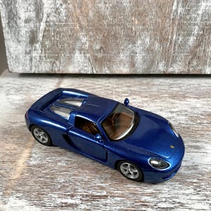 Puede incluir: Un coche de juguete azul met&aacute;lico con ruedas plateadas. El veh&iacute;culo en miniatura tiene un dise&ntilde;o elegante, con un perfil bajo y un interior detallado. El coche est&aacute; colocado sobre una superficie de madera de color claro, mostrando su color y dise&ntilde;o vibrantes.