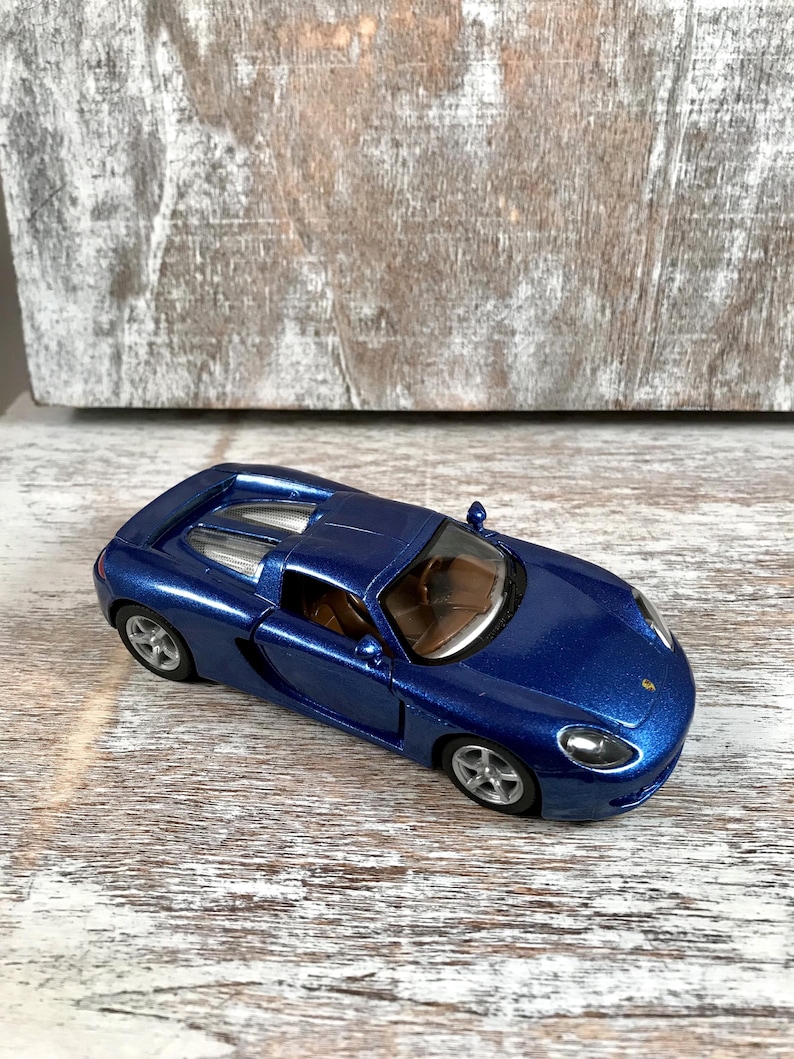 Puede incluir: Un coche de juguete azul met&aacute;lico con ruedas plateadas. El veh&iacute;culo en miniatura tiene un dise&ntilde;o elegante, con un perfil bajo y un interior detallado. El coche est&aacute; colocado sobre una superficie de madera de color claro, mostrando su color y dise&ntilde;o vibrantes.