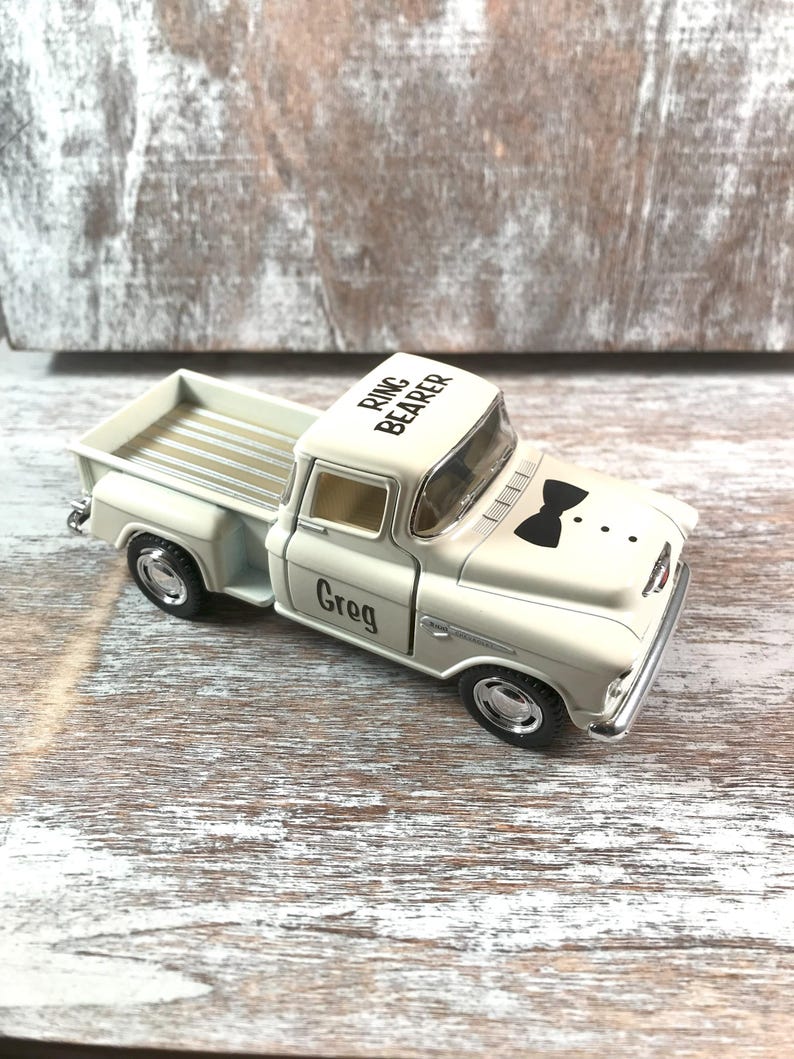 May include: A white toy truck with black wheels and chrome accents. The truck has 'Ring Bearer' printed on the roof and a black bow tie graphic on the hood. The name 'Greg' is printed on the door. The truck is sitting on a wooden surface.