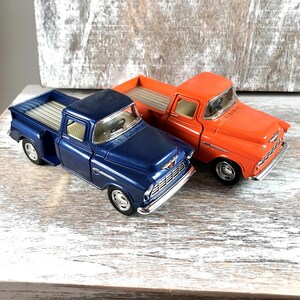 May include: Two toy pickup trucks, one in a deep blue and the other in bright orange, sit side-by-side. The trucks have silver-colored beds and chrome-colored details. The background is a weathered, light-colored wooden surface.