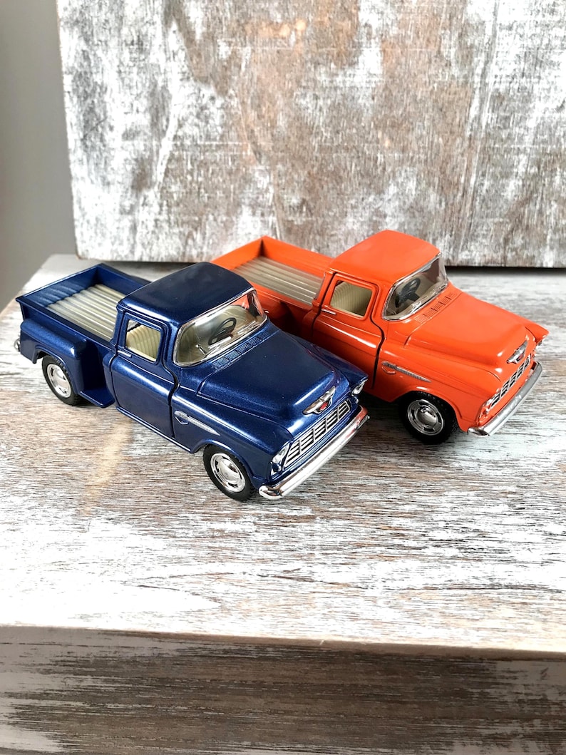May include: Two toy pickup trucks, one in a deep blue and the other in bright orange, sit side-by-side. The trucks have silver-colored beds and chrome-colored details. The background is a weathered, light-colored wooden surface.