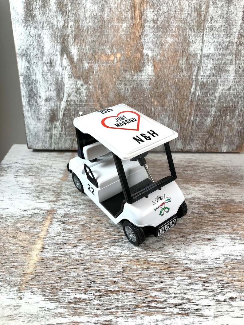 May include: White golf cart toy with a red heart that says "Just Married" and the initials "N&H" on the roof. The cart has a black steering wheel, black tires, and a black bumper. The cart has a number "22" on the side and the word "Married" on the back.
