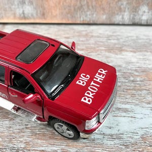 Może przedstawiać: Czerwona zabawka pick-up z białym napisem "BIG BROTHER" na masce. Samoch&oacute;d ma szyberdach, srebrne stopnie boczne i szczeg&oacute;łową przednią kratkę. Zabawkowy samoch&oacute;d stoi na drewnianej powierzchni.
