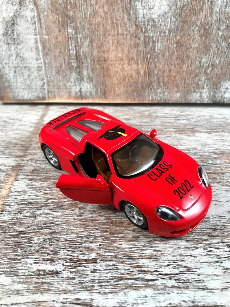 Puede incluir: Un coche de juguete rojo con el texto "CLASS OF 2022" y un gr&aacute;fico de birrete. La puerta del coche est&aacute; abierta, revelando un interior marr&oacute;n. El coche est&aacute; sobre una superficie de madera.