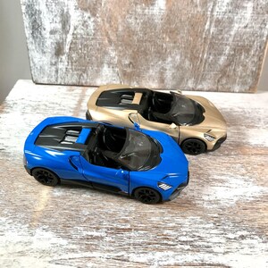 May include: Two toy sports cars, one blue and one gold, are displayed on a weathered wooden surface. The blue car is in the foreground, showcasing its sleek design. The gold car sits behind it, both with black wheels and open tops.