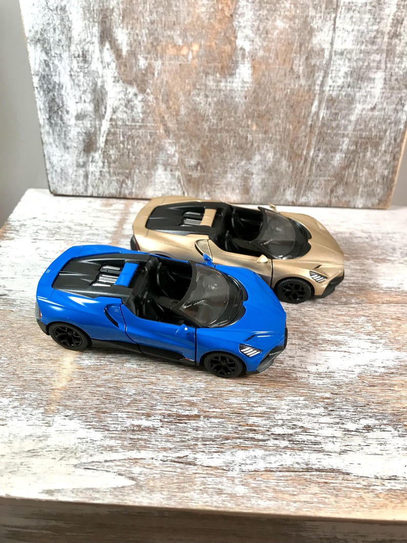 May include: Two toy sports cars, one blue and one gold, are displayed on a weathered wooden surface. The blue car is in the foreground, showcasing its sleek design. The gold car sits behind it, both with black wheels and open tops.
