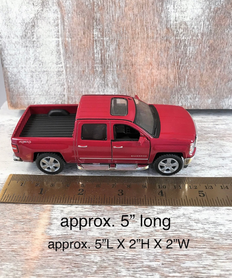 Może przedstawiać: Czerwona zabawka pick-up z czarną skrzynią i srebrnymi kołami. Ciężar&oacute;wka ma około 12,7 cm długości, 5,1 cm wysokości i 5,1 cm szerokości. Ciężar&oacute;wka ma szyberdach, a na boku widnieje napis "Silverado".