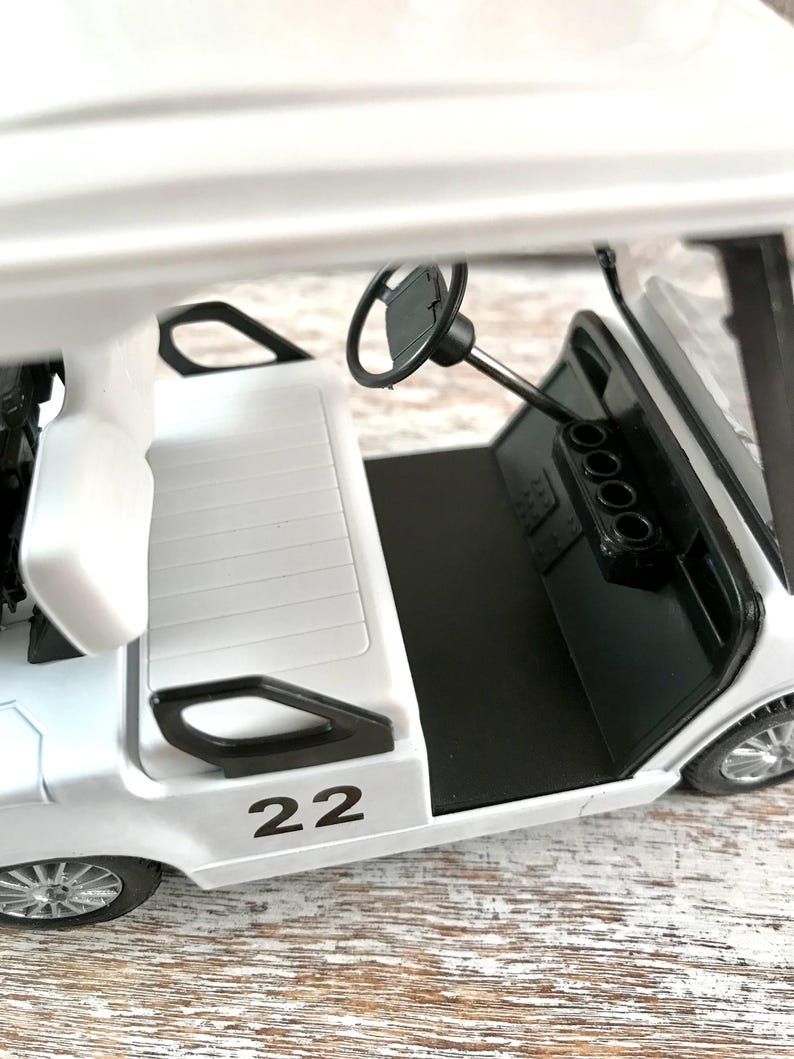 May include: A white golf cart toy with black accents, including the steering wheel and dashboard. The number "22" is printed in black on the side. The cart has a black floor and silver wheels.