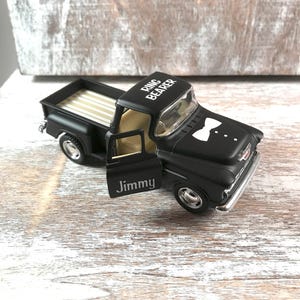 May include: A black toy pickup truck with the words "Ring Bearer" on the roof and "Jimmy" on the door. The truck has a white bow tie design on the hood and a tan interior.