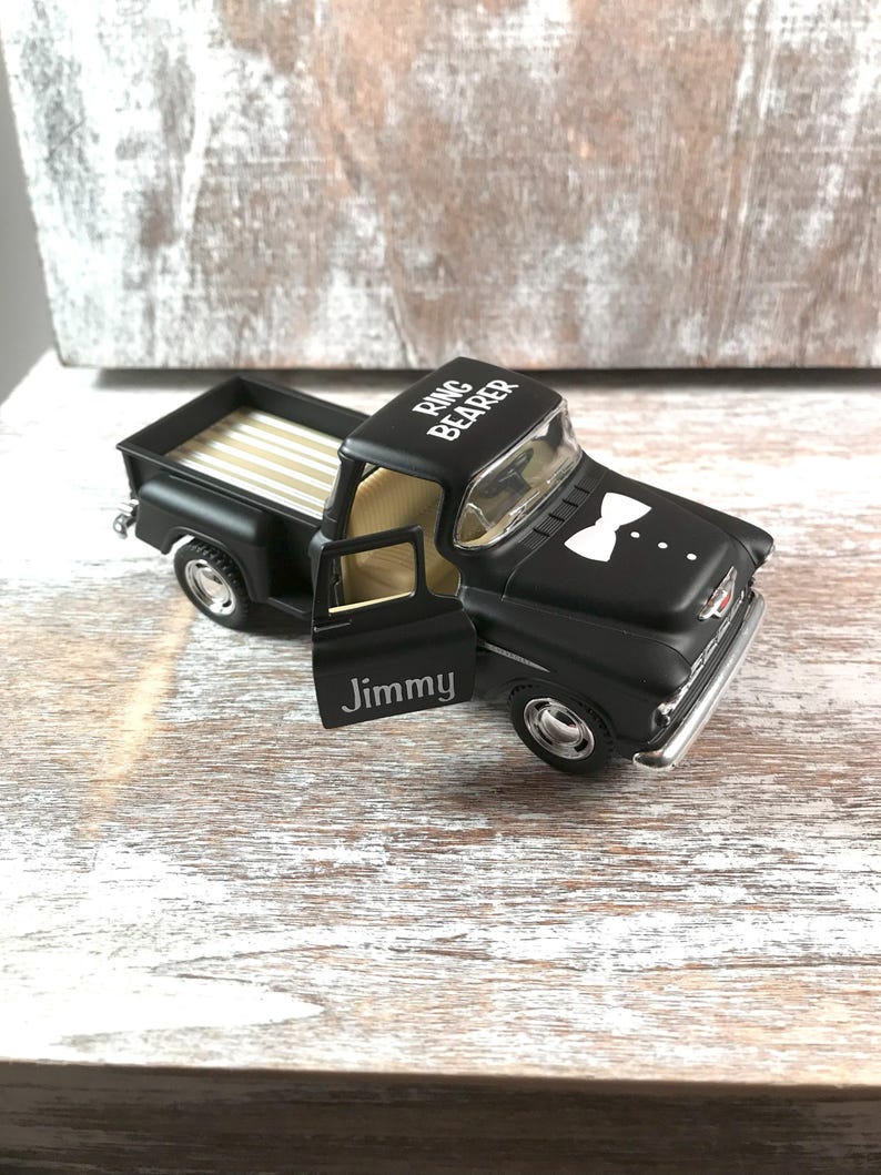 May include: A black toy pickup truck with the words "Ring Bearer" on the roof and "Jimmy" on the door. The truck has a white bow tie design on the hood and a tan interior.