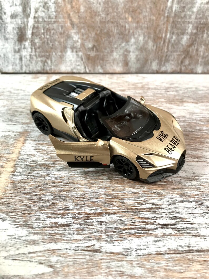 May include: A gold-colored toy convertible car with black wheels and interior. The car's door is open, revealing the name "KYLE" printed on the side. The words "RING BEARER" are printed on the front.