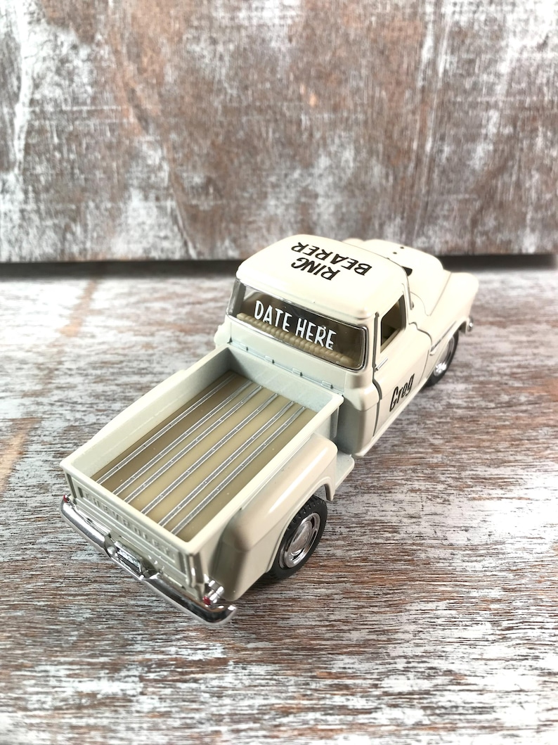 May include: A cream-colored toy pickup truck with the words "DATE HERE" on the windshield and "RING BEARER" on the roof. The truck bed is beige with wooden slats. The truck has chrome details.