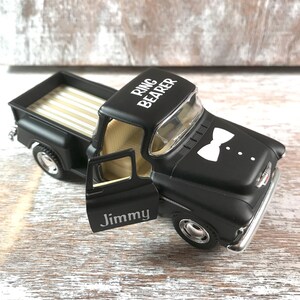 May include: A black toy pickup truck with the words "Ring Bearer" on the roof and a white bow tie graphic on the hood. The door is open, revealing the name "Jimmy". The truck bed has a gold and white striped pattern.