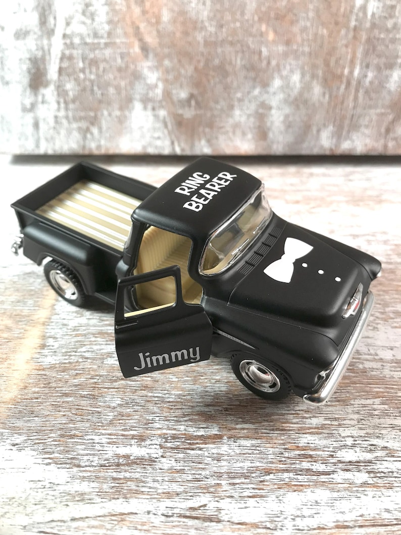 May include: A black toy pickup truck with the words "Ring Bearer" on the roof and a white bow tie graphic on the hood. The door is open, revealing the name "Jimmy". The truck bed has a gold and white striped pattern.