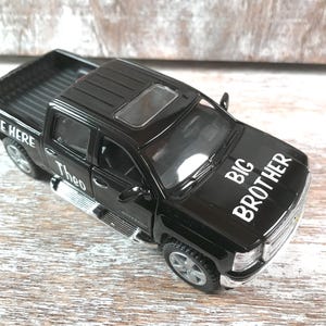 Może przedstawiać: Czarny zabawkowy pickup z białym napisem "BIG BROTHER" na masce. Samoch&oacute;d ma szyberdach i imię "Theo" z boku. Samoch&oacute;d jest modelem Chevrolet Silverado.