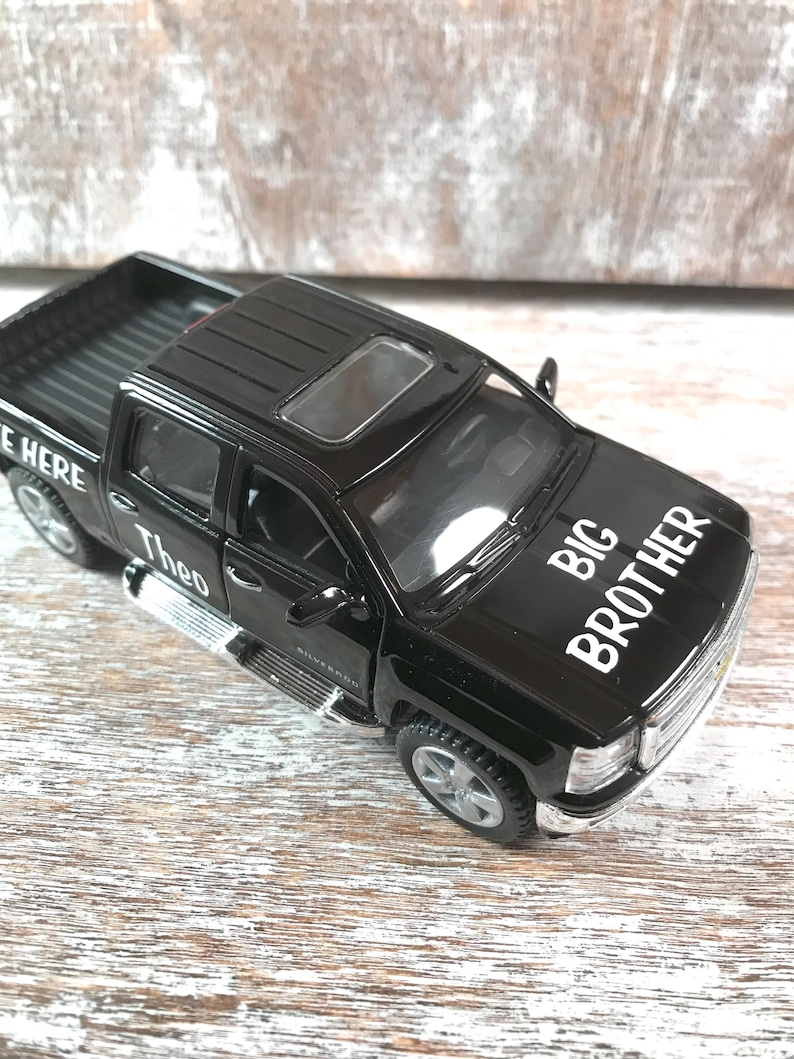 Może przedstawiać: Czarny zabawkowy pickup z białym napisem "BIG BROTHER" na masce. Samoch&oacute;d ma szyberdach i imię "Theo" z boku. Samoch&oacute;d jest modelem Chevrolet Silverado.