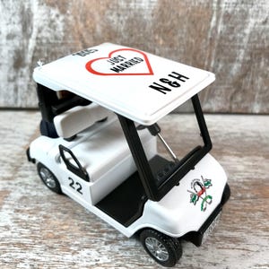 May include: White golf cart toy with a red heart that says "Just Married" and the initials "N&H" on the roof. The cart has a black steering wheel, black seats, and black wheels. The cart has a number "22" on the side and a golf club logo on the back.