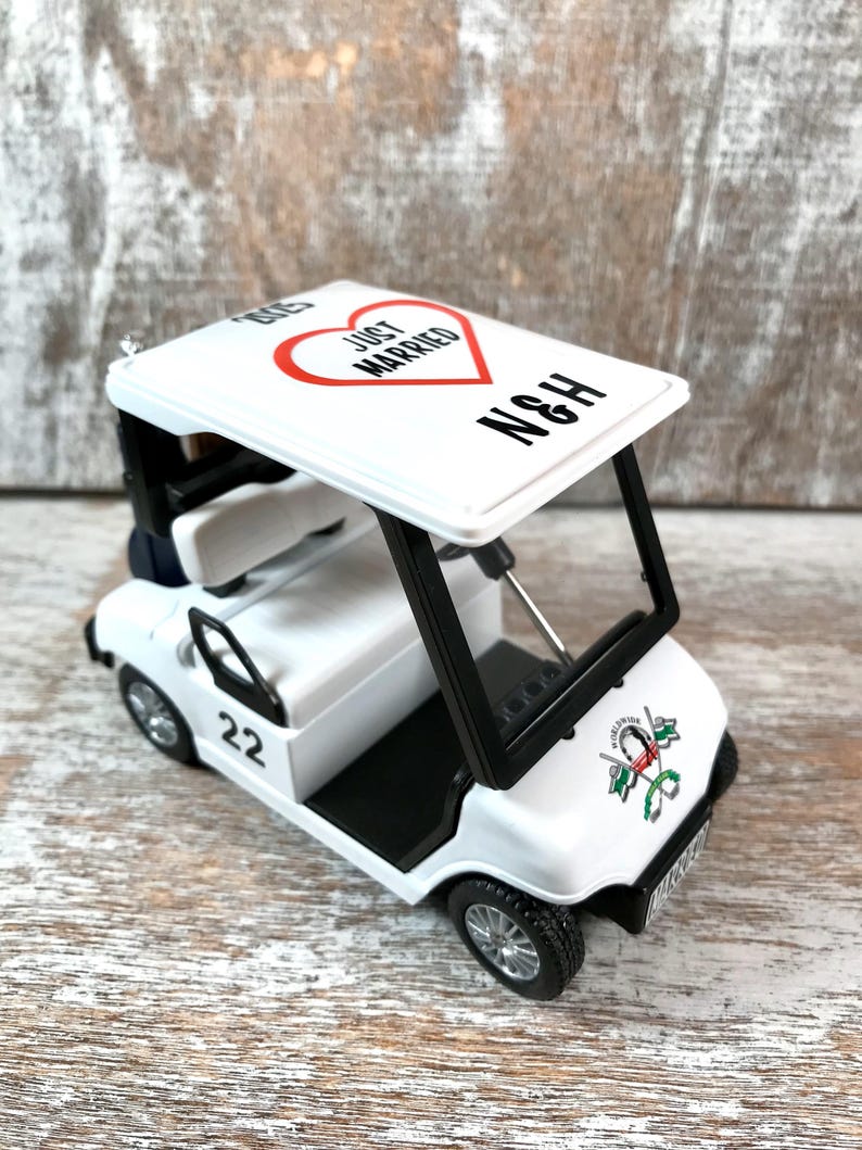 May include: White golf cart toy with a red heart that says "Just Married" and the initials "N&H" on the roof. The cart has a black steering wheel, black seats, and black wheels. The cart has a number "22" on the side and a golf club logo on the back.
