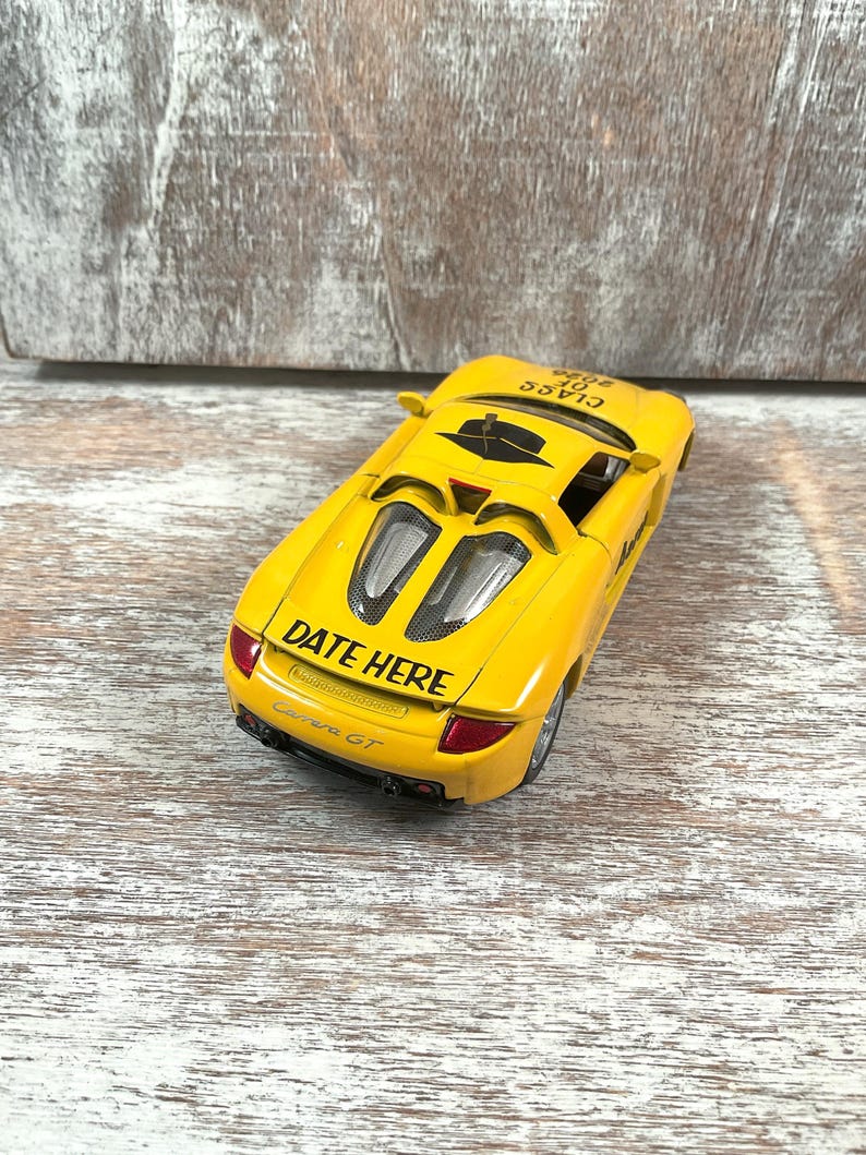 Puede incluir: Un coche de juguete amarillo con el texto "DATE HERE" y "Carrera GT" impreso en la parte trasera. El coche tiene detalles en negro y un interior negro. Las palabras "CLASS OF 2006" est&aacute;n impresas en la parte superior del coche.