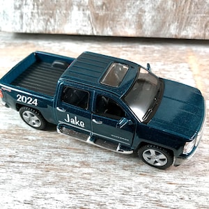 Może przedstawiać: Tealowa zabawka pick-up z napisem "Jake" i rokiem "2024". Samoch&oacute;d ma szyberdach i srebrne akcenty. Widoczne jest r&oacute;wnież słowo "Silverado".