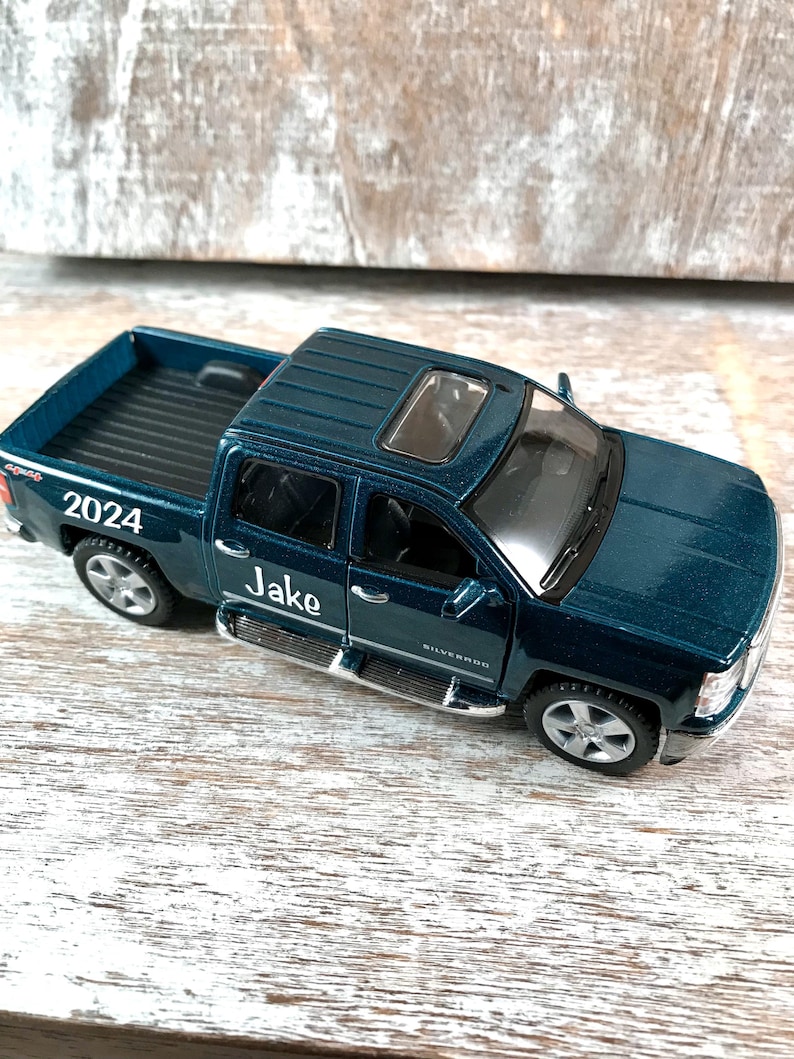 Może przedstawiać: Tealowa zabawka pick-up z napisem "Jake" i rokiem "2024". Samoch&oacute;d ma szyberdach i srebrne akcenty. Widoczne jest r&oacute;wnież słowo "Silverado".