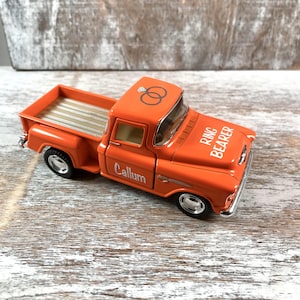 May include: An orange toy pickup truck with the words "Ring Bearer" and "Callum" printed on it. The truck has a silver bed and wheels, and a graphic of two wedding rings on the roof.